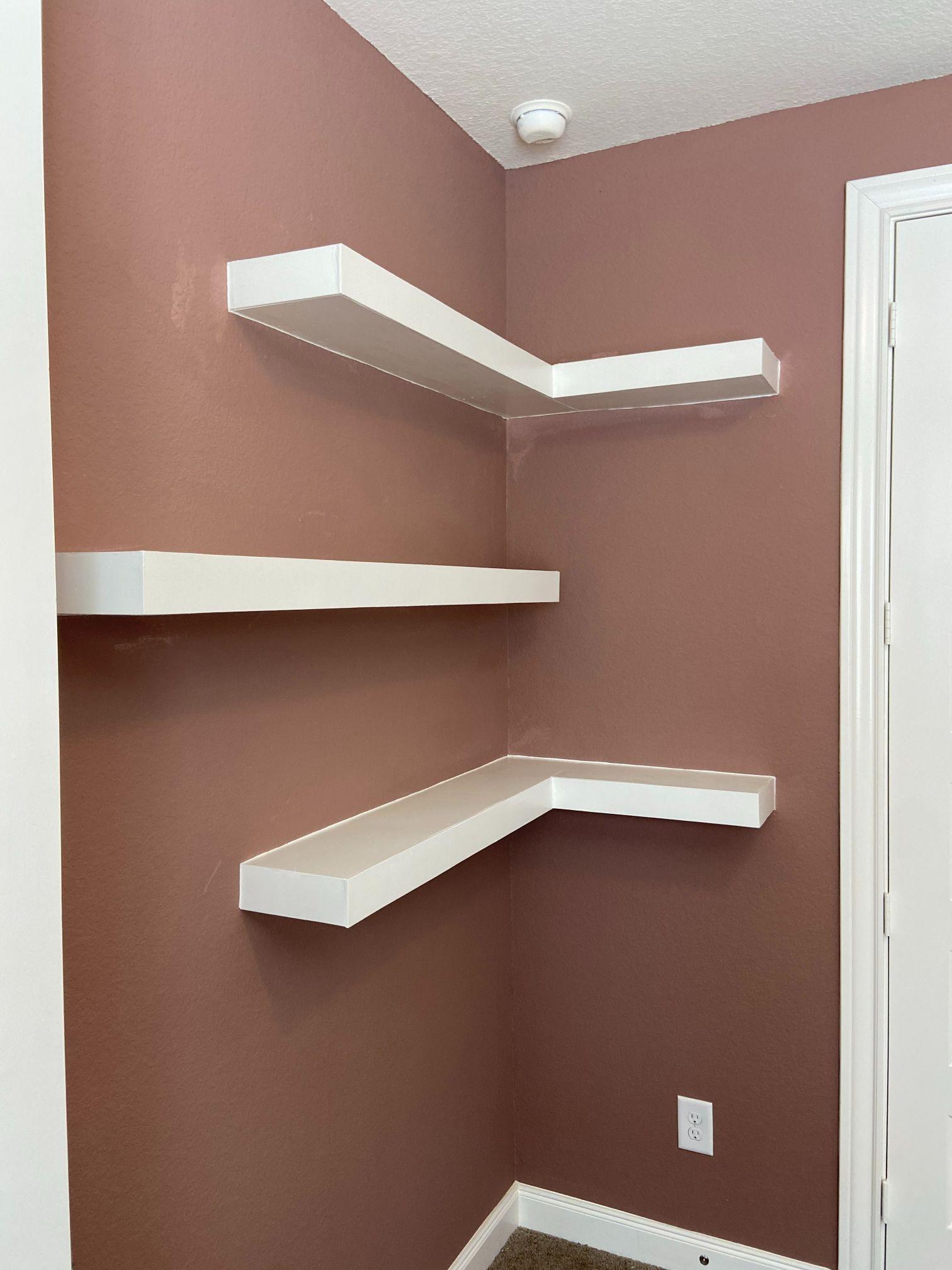Empty mauve corner with three white floating shelves and a small wall outlet.