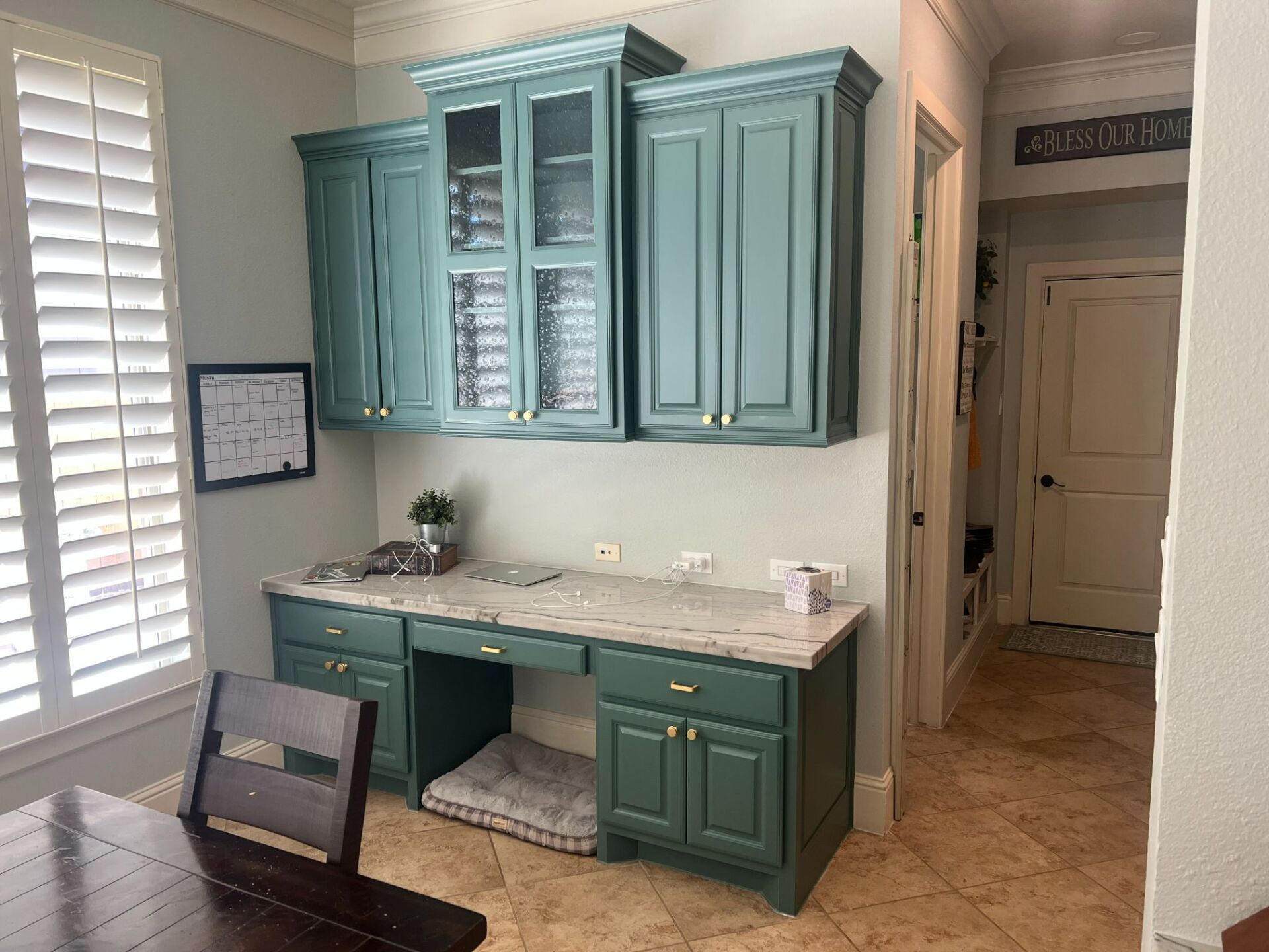Teal built-in desk with cabinets and marble top in a bright room with shutters and tan tile floor