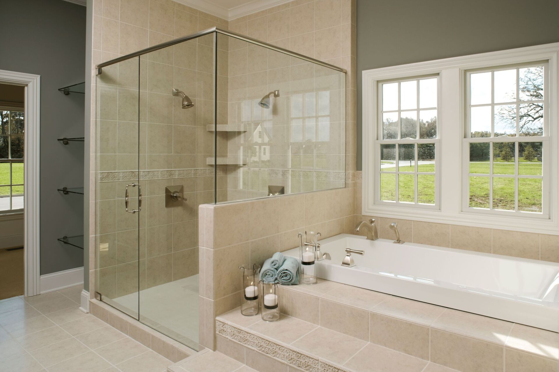 Bright bathroom with glass walk-in shower, soaking tub, beige tile, and two windows overlooking a green yard