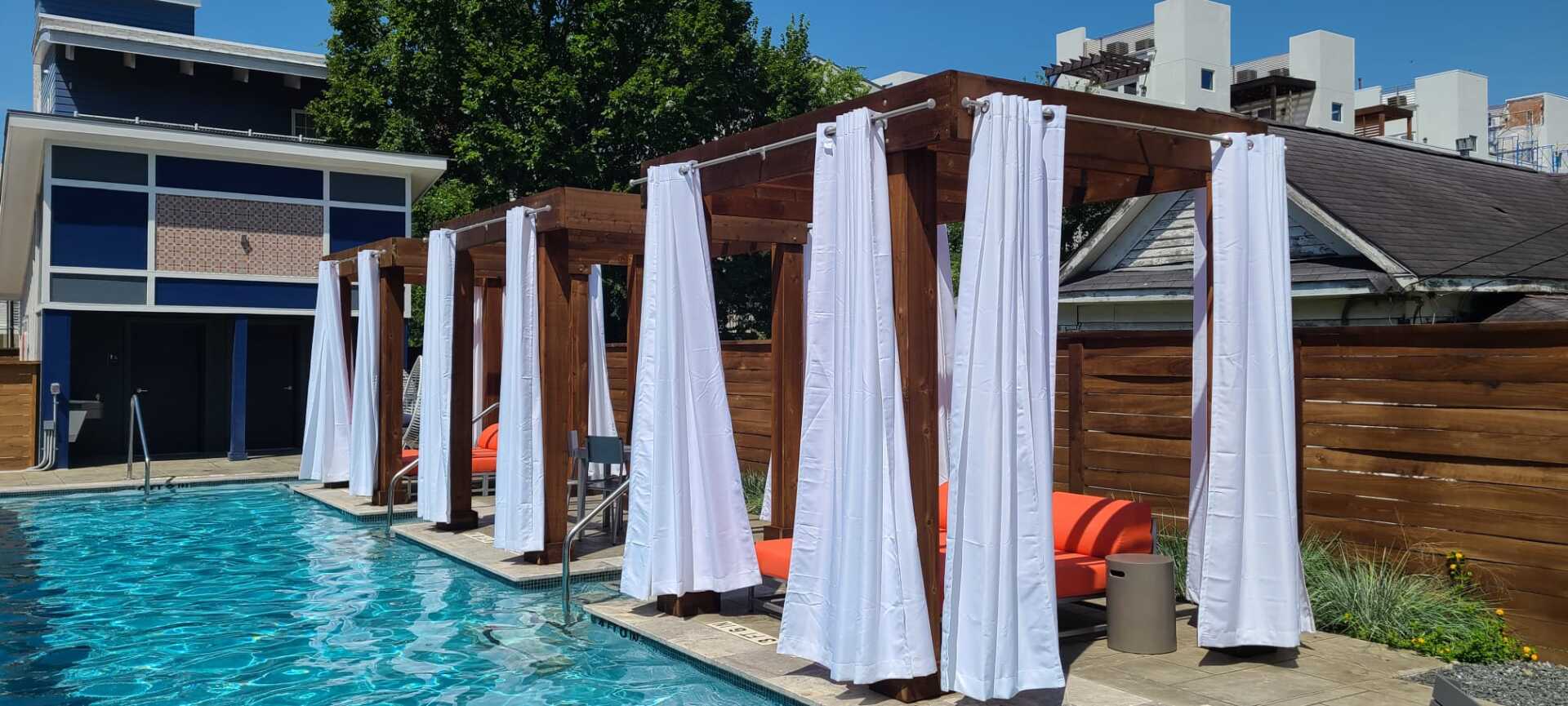 Poolside cabanas with white curtains beside a turquoise swimming pool and wooden deck