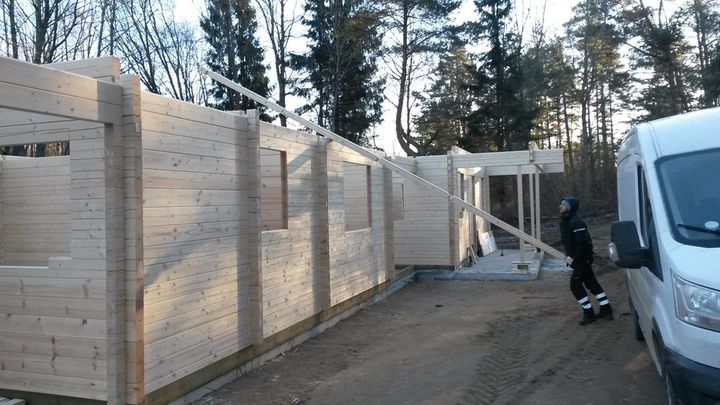Construction site with workers framing a large wooden wall beside a parked white van and trees.