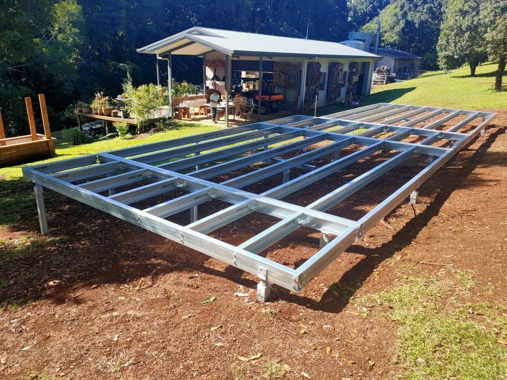 Steel foundation frame under construction on a dirt lot beside a small shed in a sunny yard