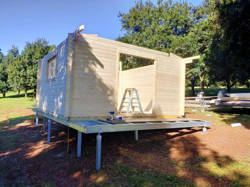 Partially built wooden cabin on raised posts, with ladder and open doorway in a sunny yard