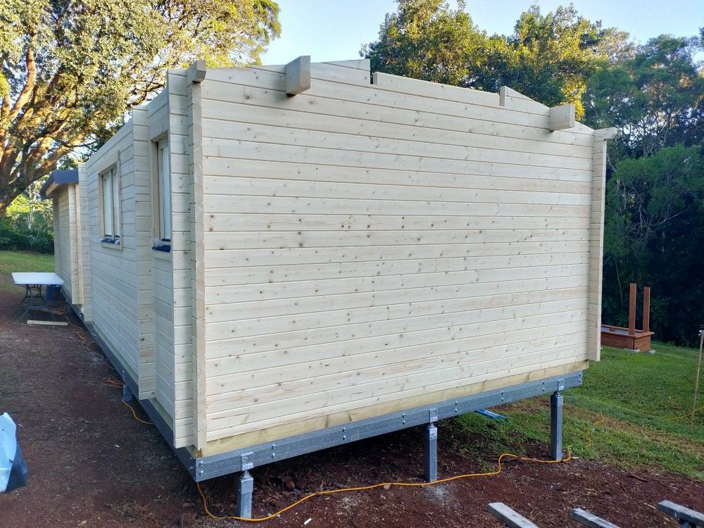 Small white wooden cabin on metal supports in a grassy outdoor area with trees nearby