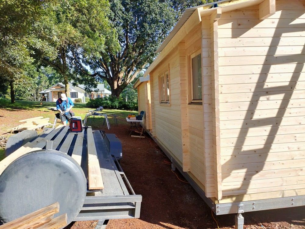 Wooden shed beside a patio table and benches in a sunny backyard with trees and a person in the distance