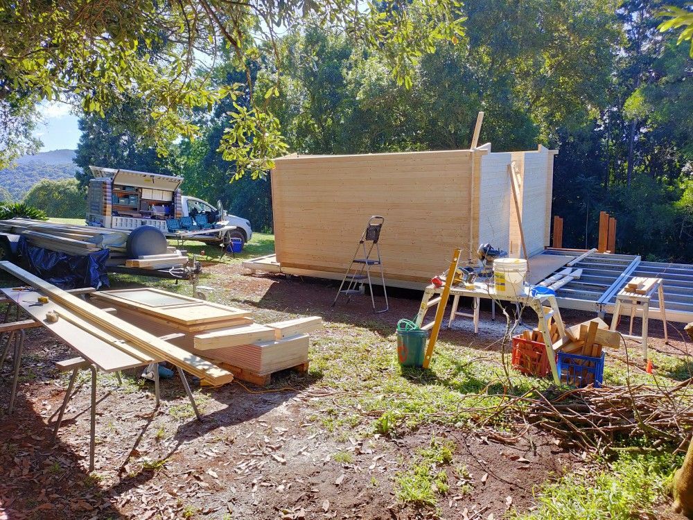 Construction site with a wooden shed frame, lumber piles, tools, and a ladder in a sunny yard