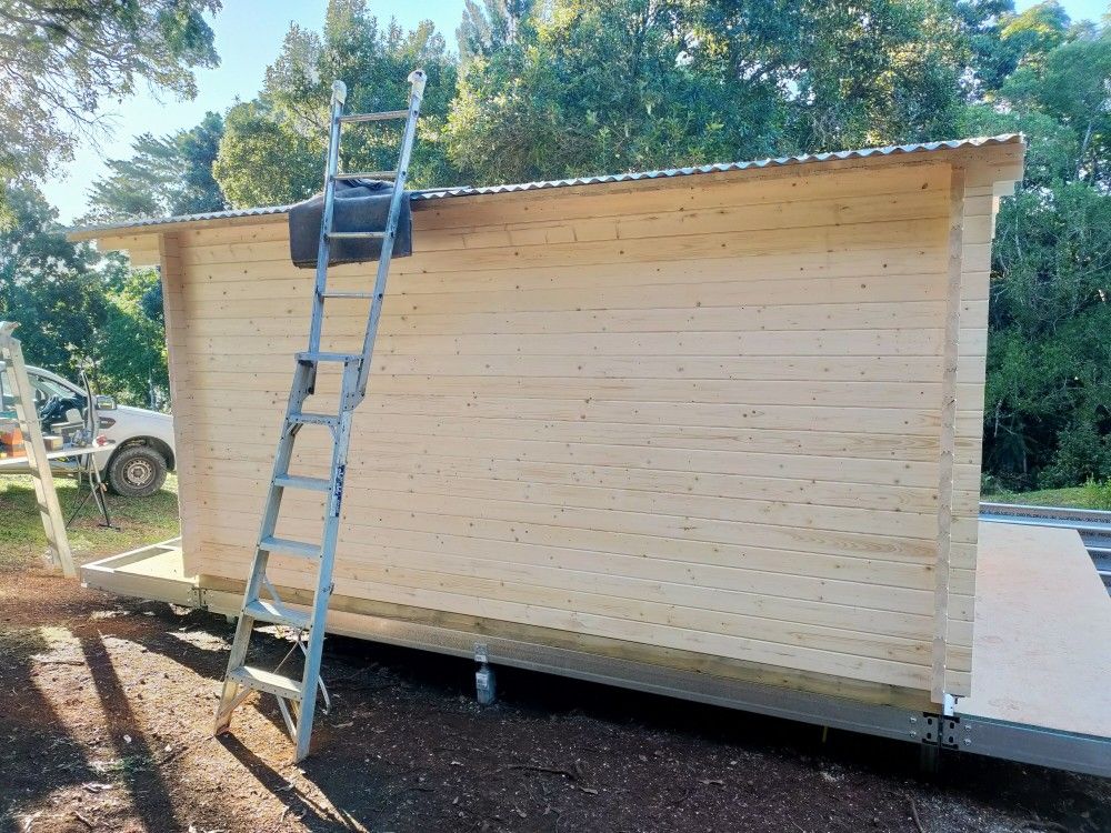 Wooden cabin under construction with a ladder leaning against the roof outdoors.
