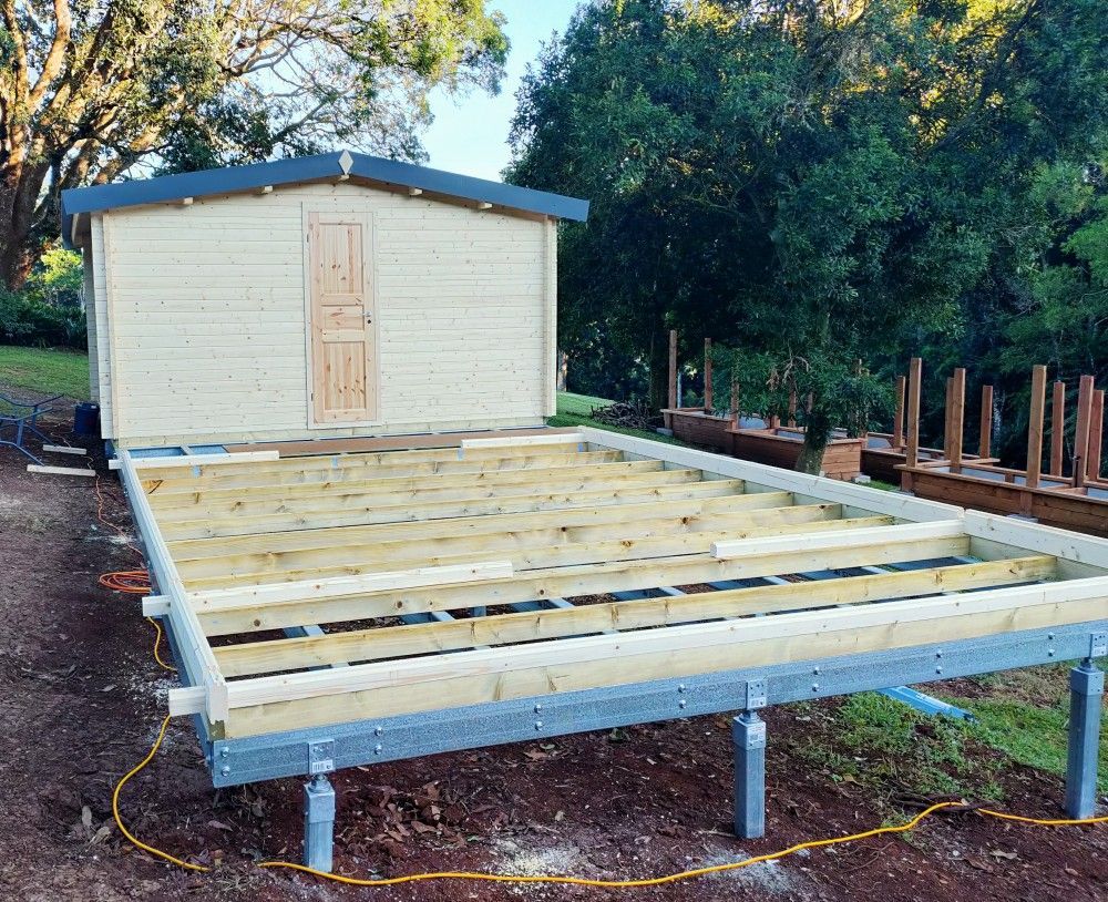 Raised wooden deck frame under construction beside a small beige shed in a wooded yard