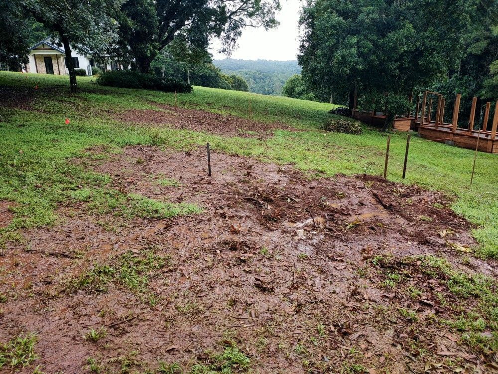 Muddy grassy yard with a large disturbed patch, trees, and a house in the background