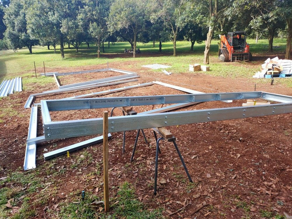 Steel frame beams laid on supports in a rural yard, with an orange tractor in the background.