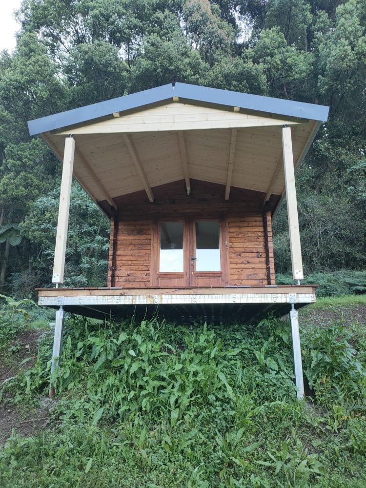 Small wooden cabin with a covered porch on stilts, surrounded by lush green forest.
