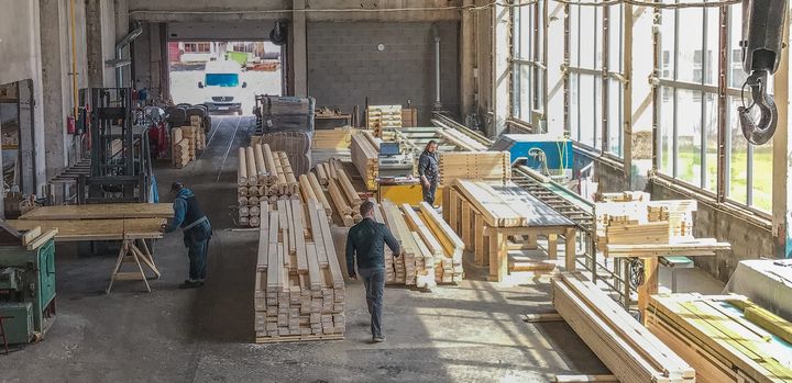Workers moving lumber in a bright industrial wood workshop with stacks of timber and machinery