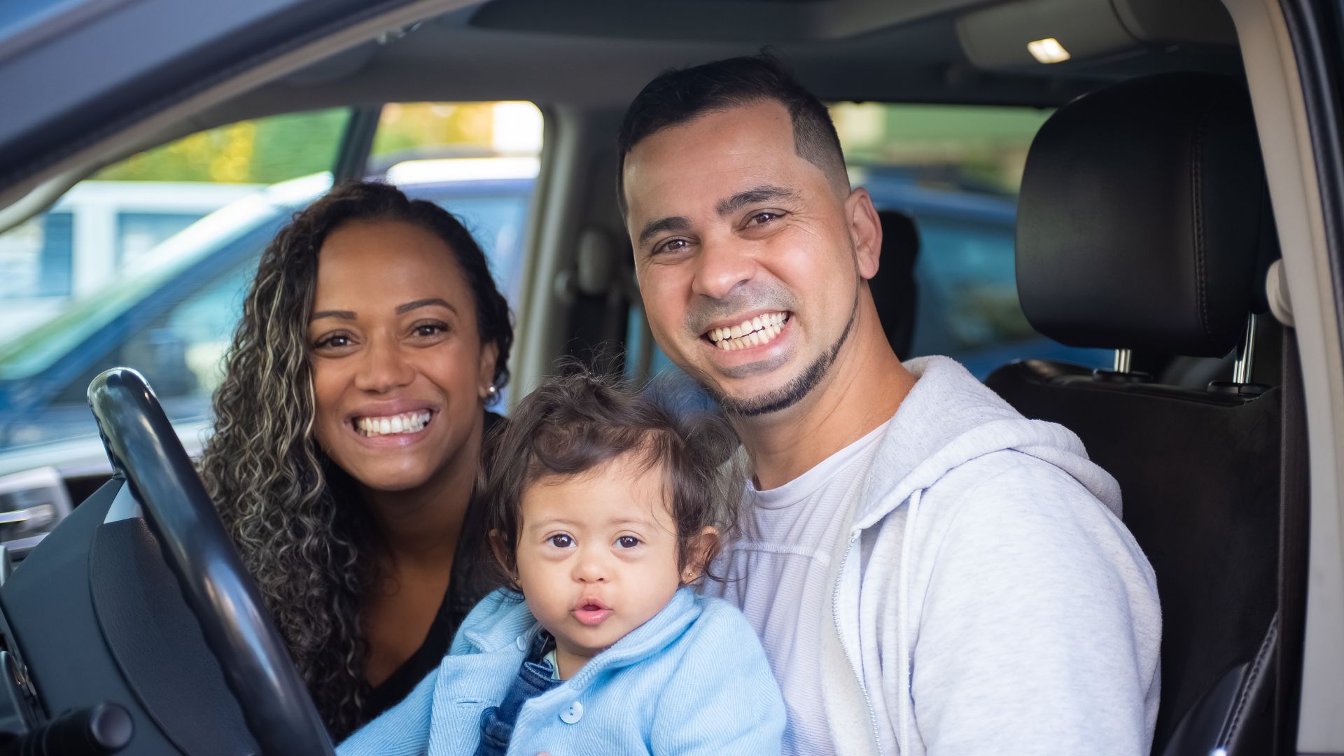 Smiling family sitting together inside a car, with a baby in the front seat.