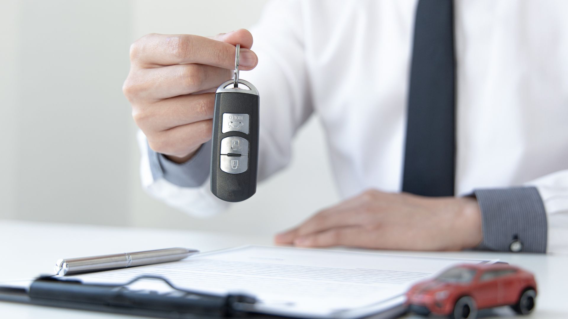 Hand holding a car key fob over a desk with papers, a notebook, and a red toy car