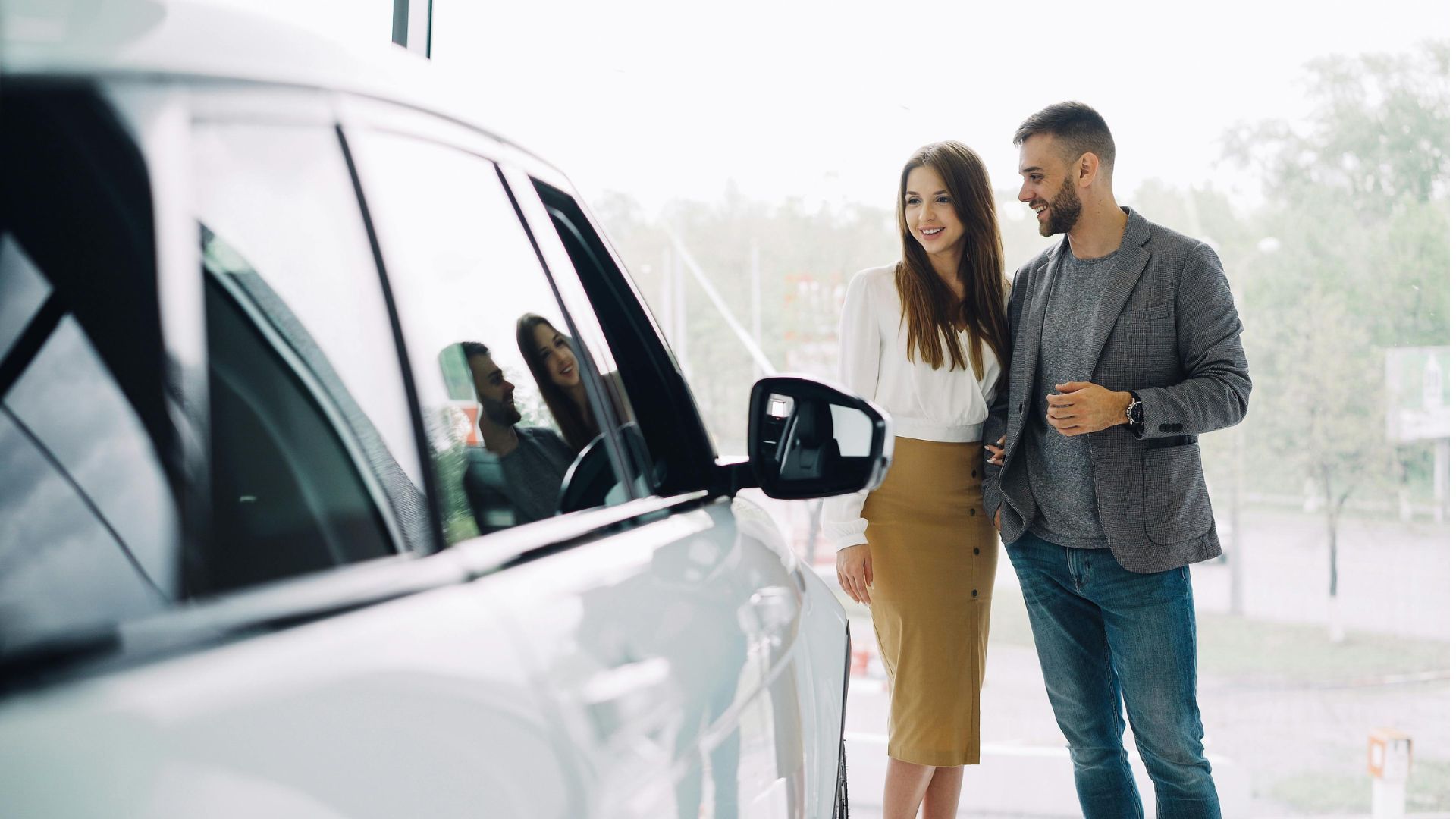 Couple standing beside a white car in a bright outdoor setting, looking at the vehicle and smiling.