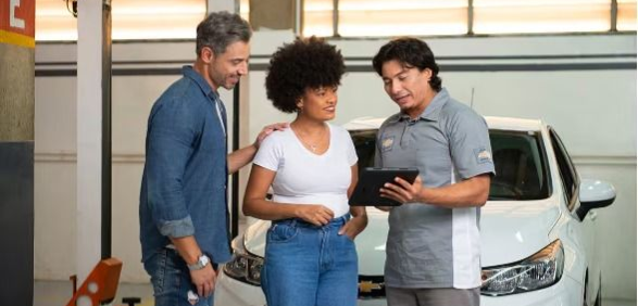Three people stand beside a white car, looking at a tablet in a garage or showroom.