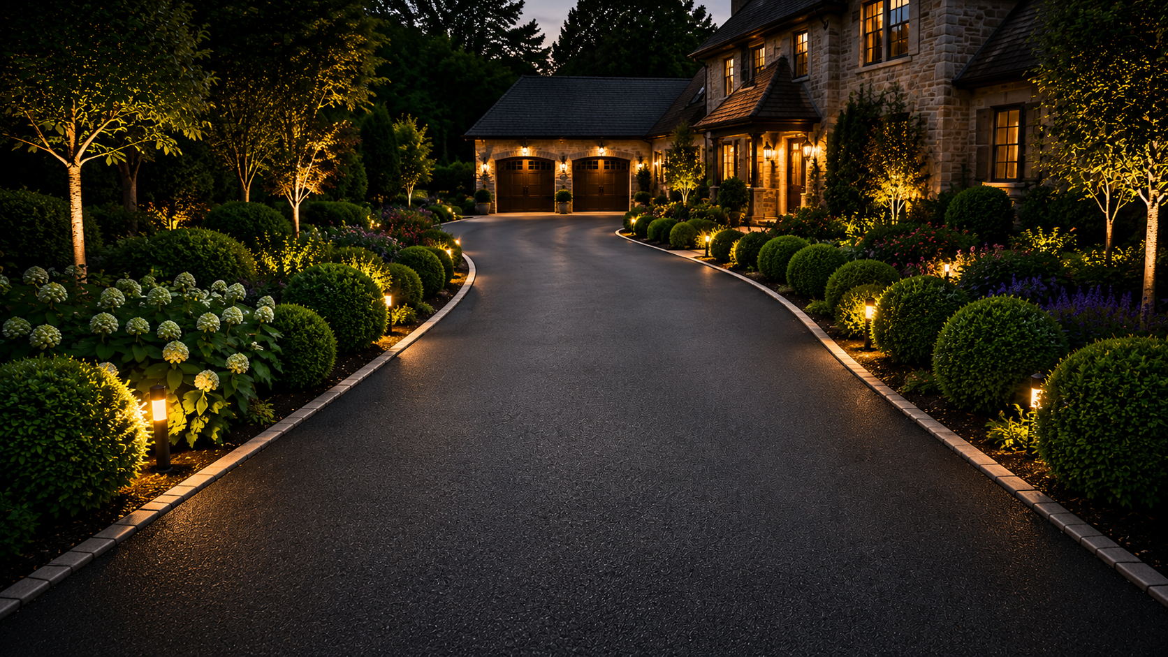 Dusk view of a curved driveway leading to a warmly lit stone house with landscaped shrubs and trees