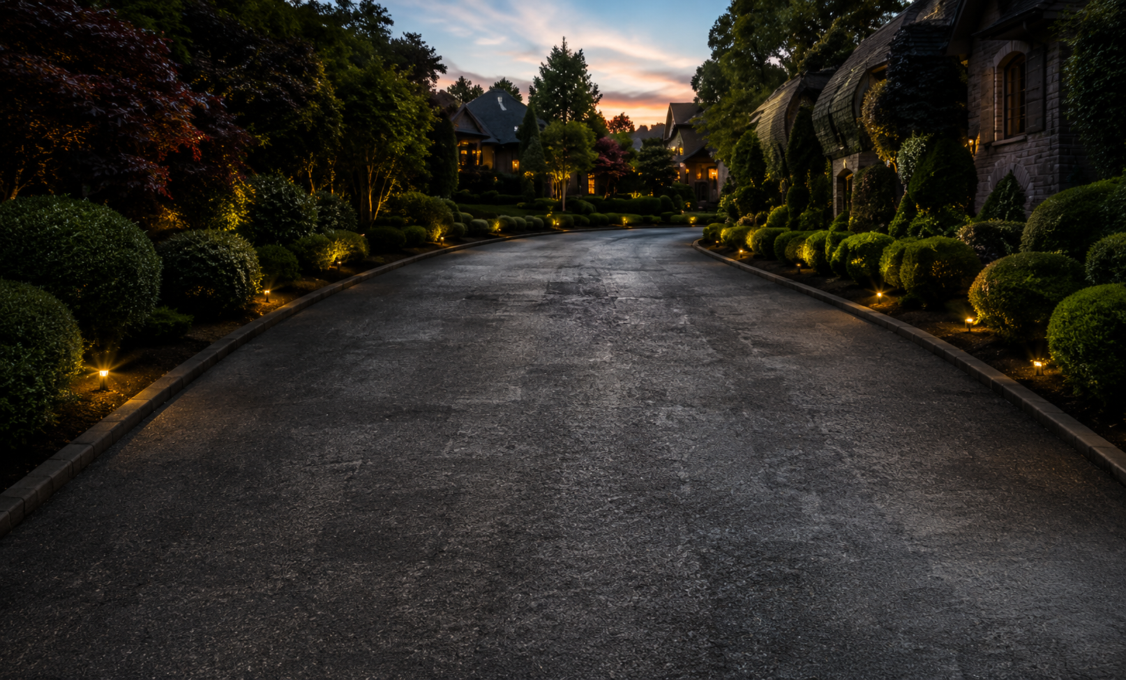 Curved asphalt road lined with hedges and flowers at sunset, with houses in the background