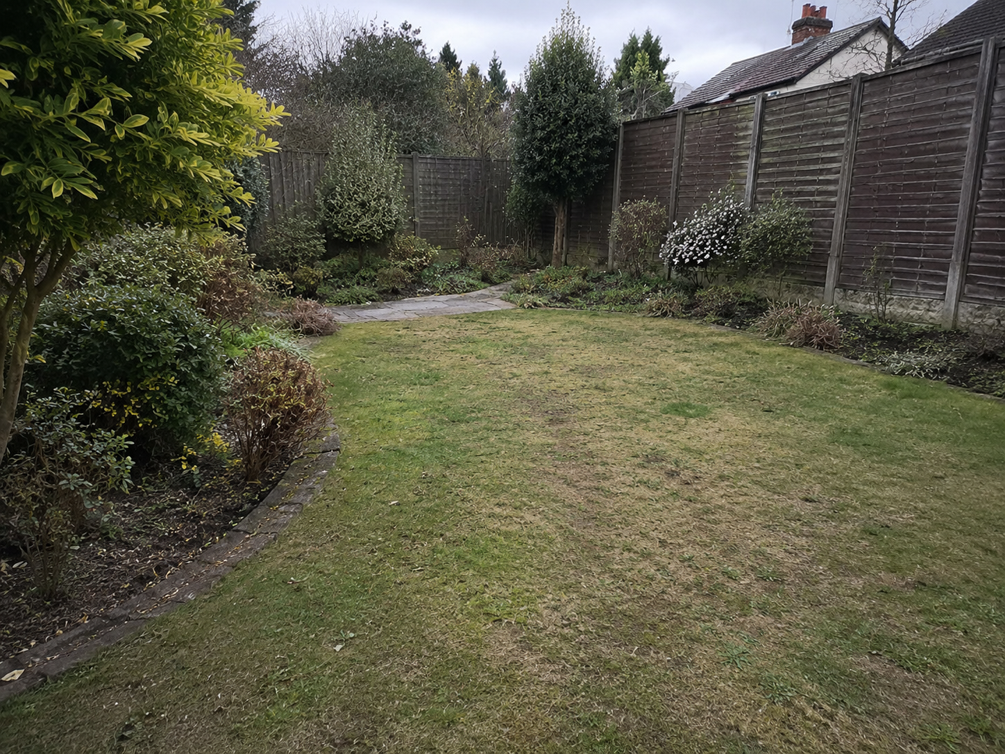 Backyard lawn bordered by shrubs, trees, and a wooden fence on a cloudy day