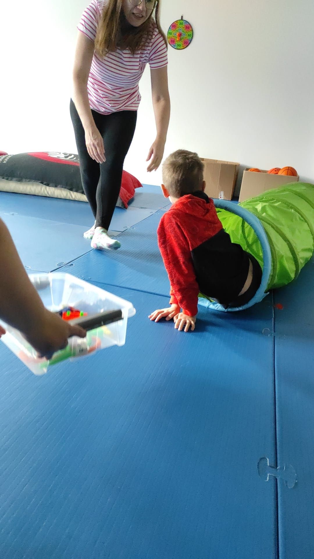 A person helps a child crawl through a green fabric tunnel in a room with blue floor mats and a toy box.