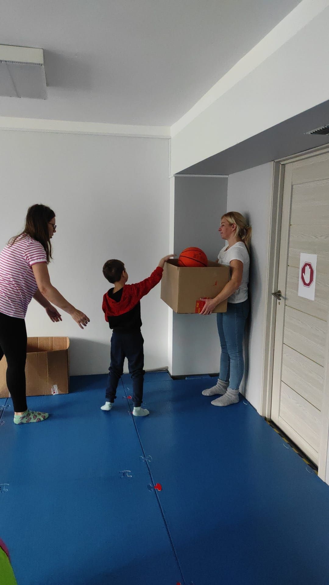 Two people help a child place an orange basketball into a cardboard box held by someone standing near a door.
