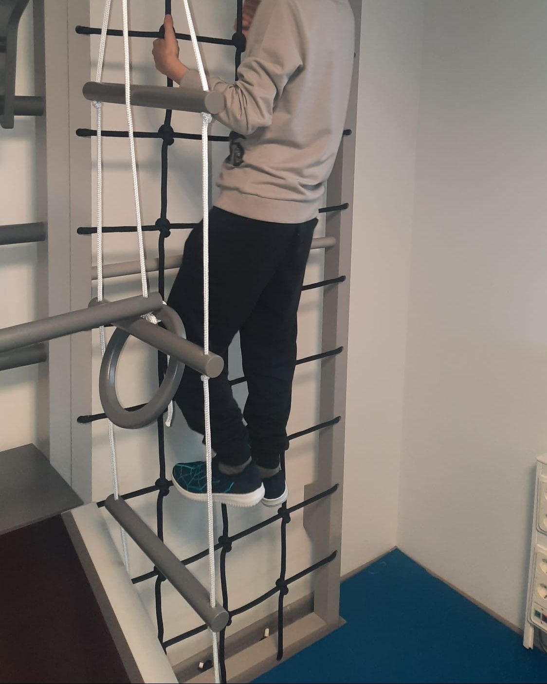 A person wearing a grey sweater and black joggers climbs a wooden wall ladder and rope net structure in a gym.