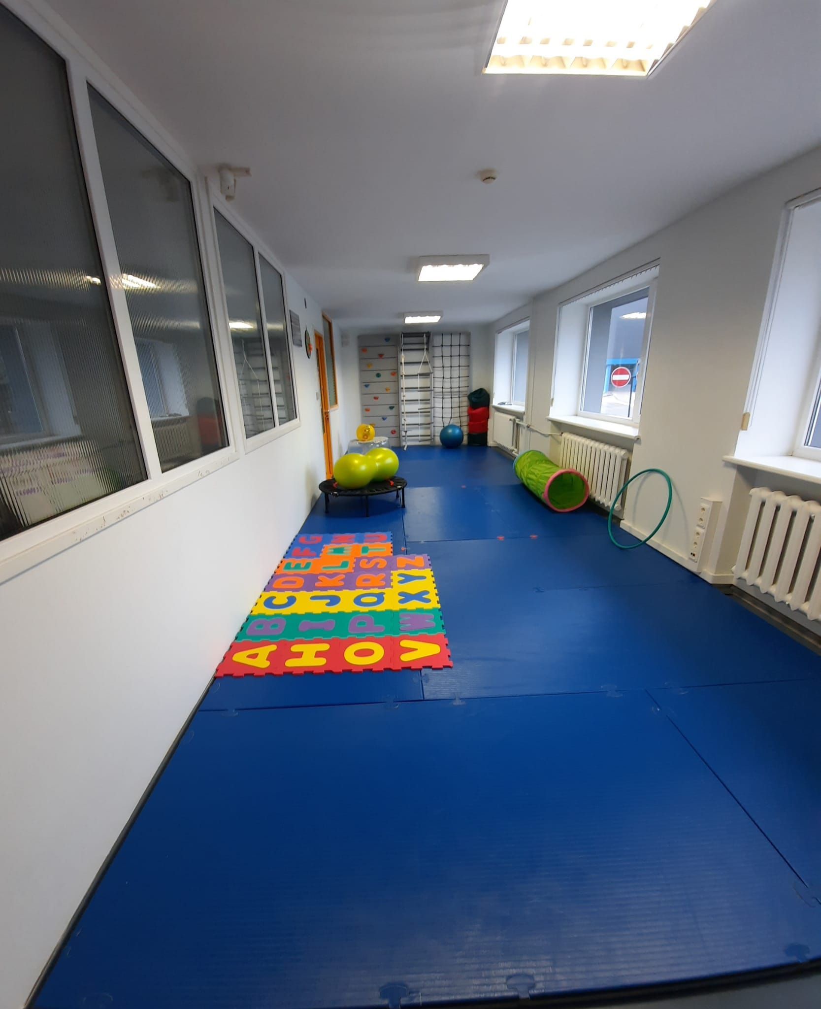 A long, narrow pediatric therapy room with blue floor mats, colorful alphabet puzzle tiles, a play tunnel, and gym balls.