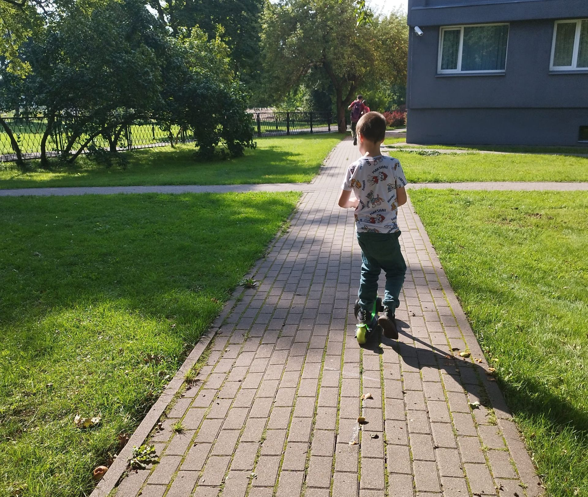 A child riding a scooter on a brick walkway outdoors next to a grassy area and a building.