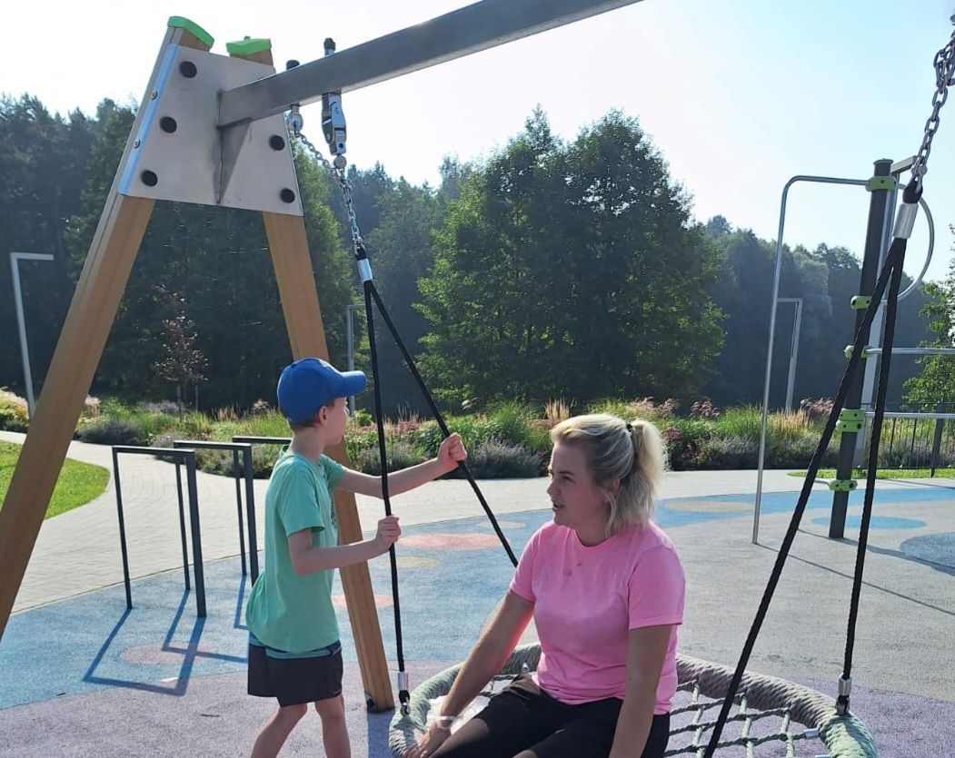 A child and a person sit together on a saucer swing at an outdoor playground on a sunny day.