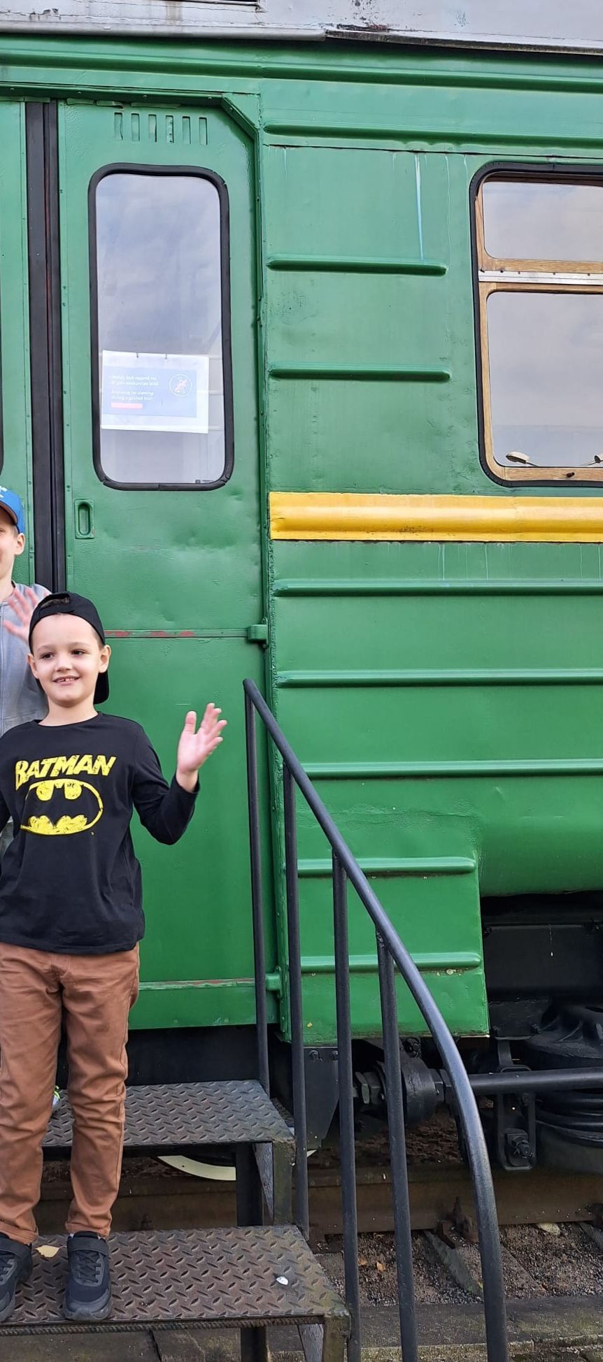 A child wearing a Batman shirt and baseball cap stands on the metal stairs of a green vintage train carriage.