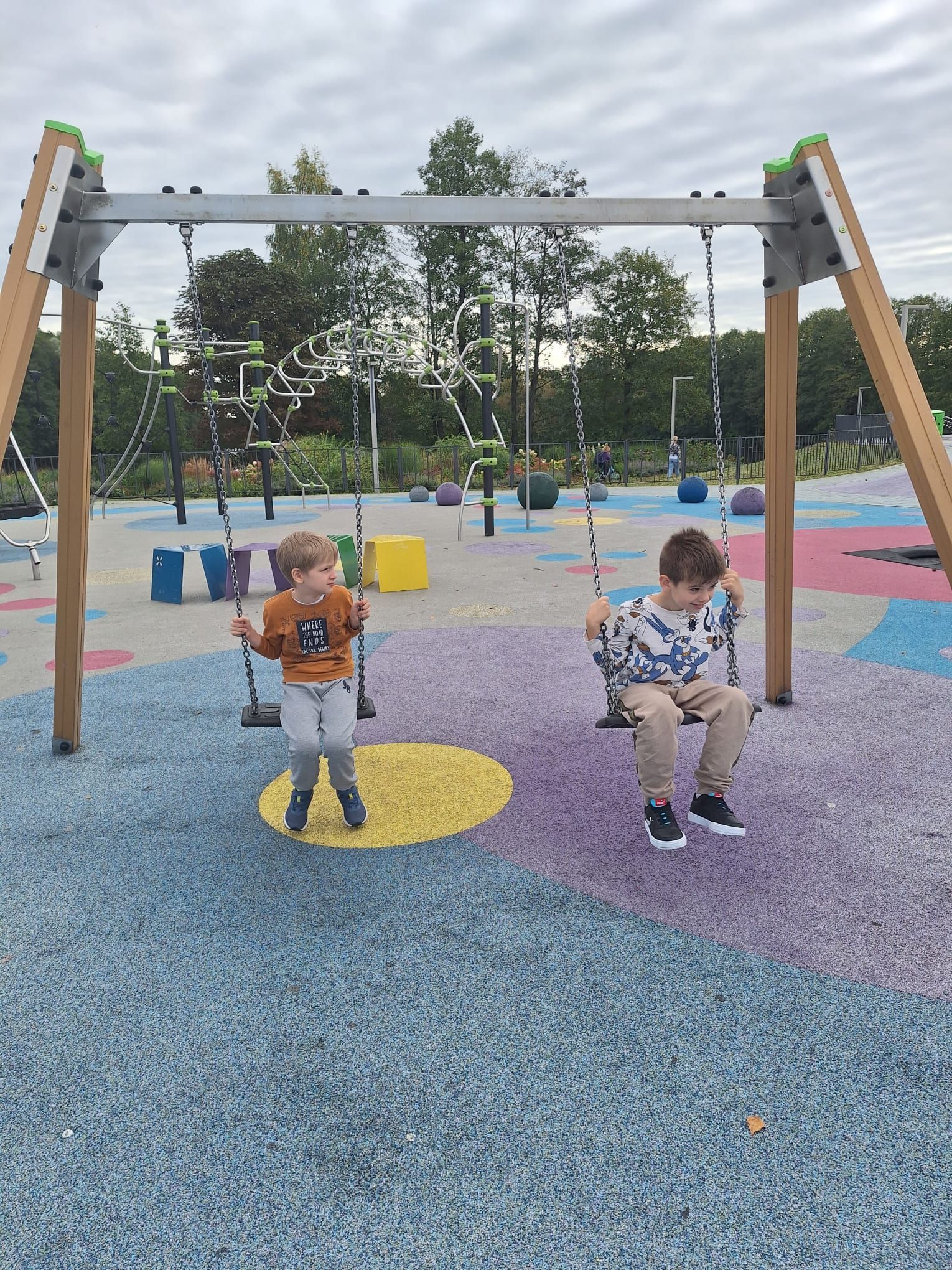 Two children sit on a swing set at a colorful, paved outdoor playground with trees in the background.