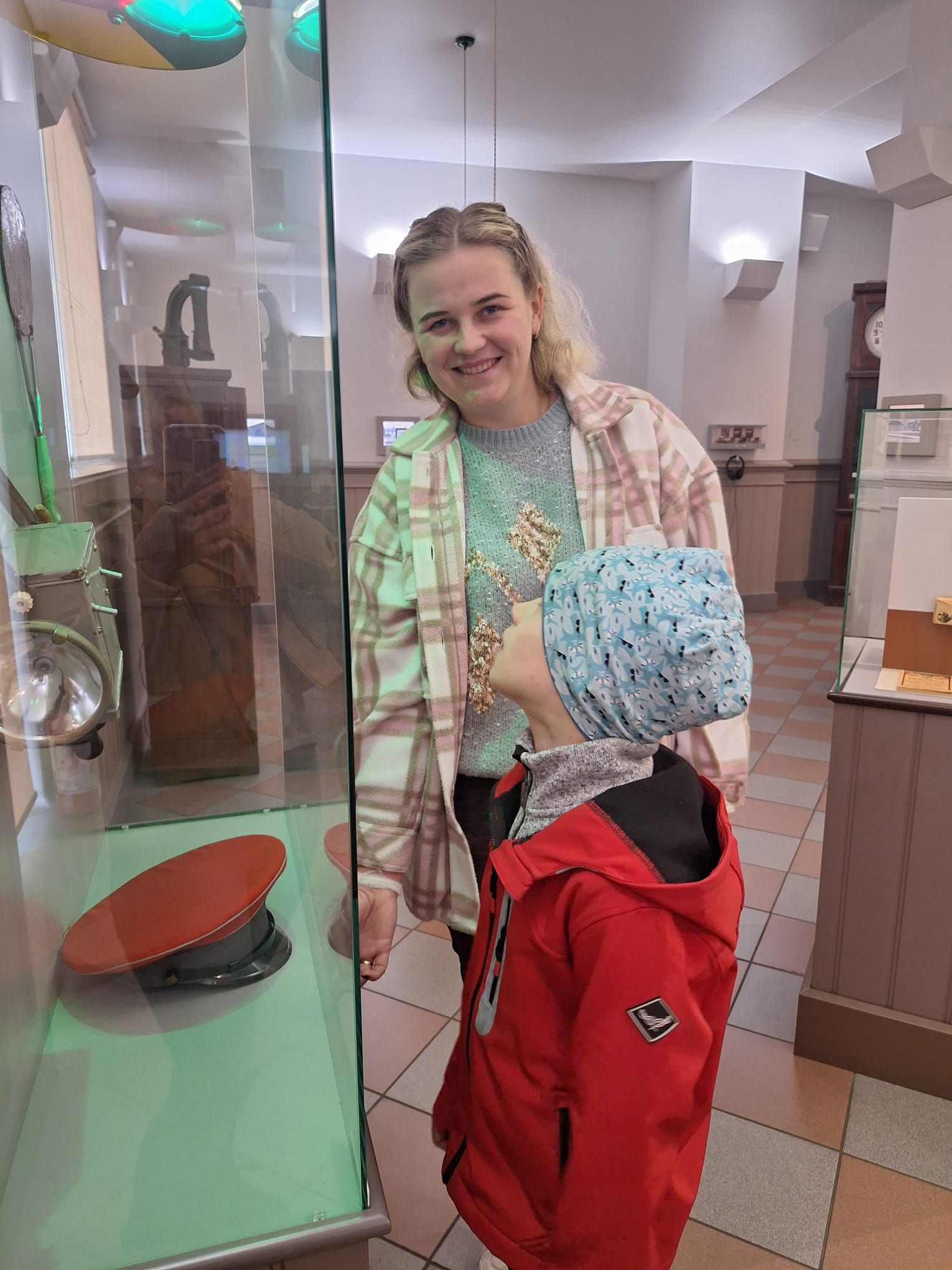 A person wearing a plaid shirt and a child in a red jacket look at a military cap displayed in a glass museum case.