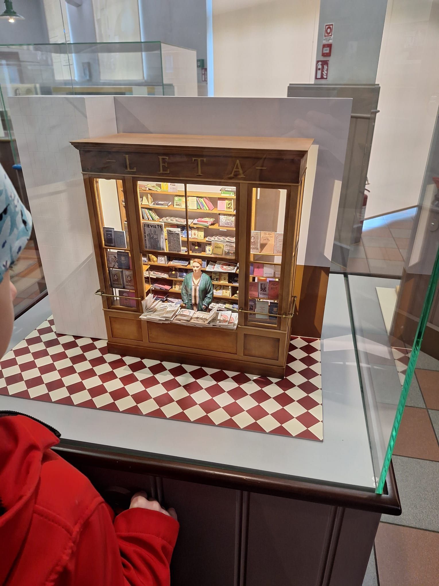 A miniature, detailed store model with a checkered floor, seen from the perspective of a visitor in a red jacket.