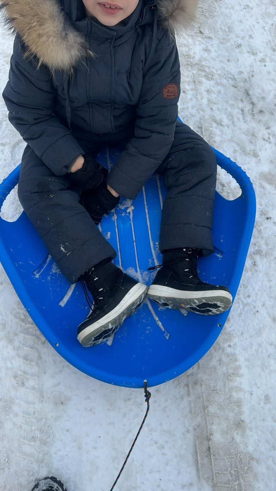 A person wearing a dark winter coat with a fur-trimmed hood and black boots sitting on a blue saucer sled in the snow.