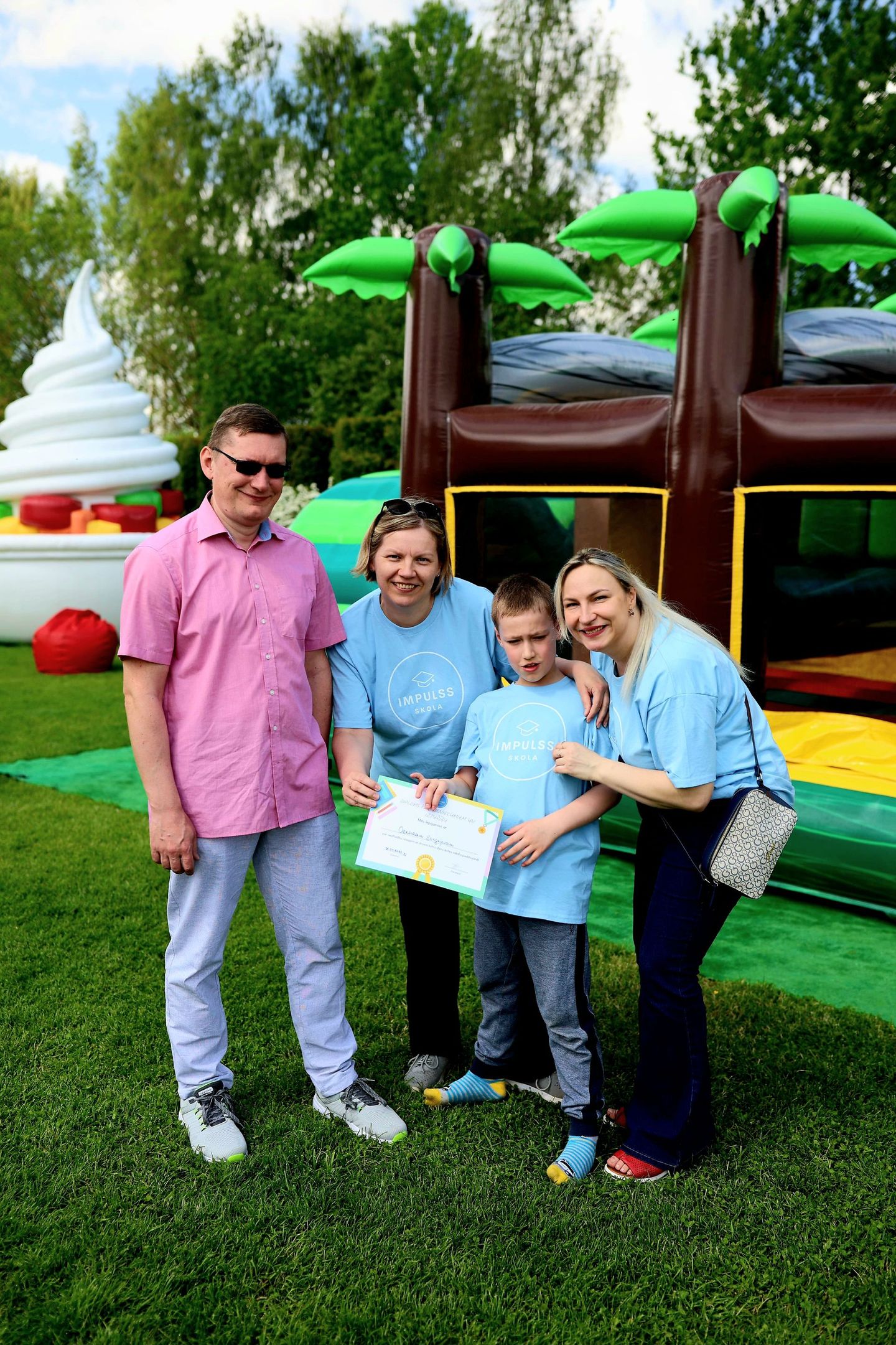 A family of four stands outdoors in front of a bouncy castle, with the child holding a certificate.