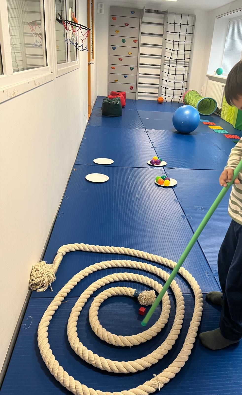 A child uses a stick to guide a small ball through a spiral rope maze on a blue floor in an indoor activity room.