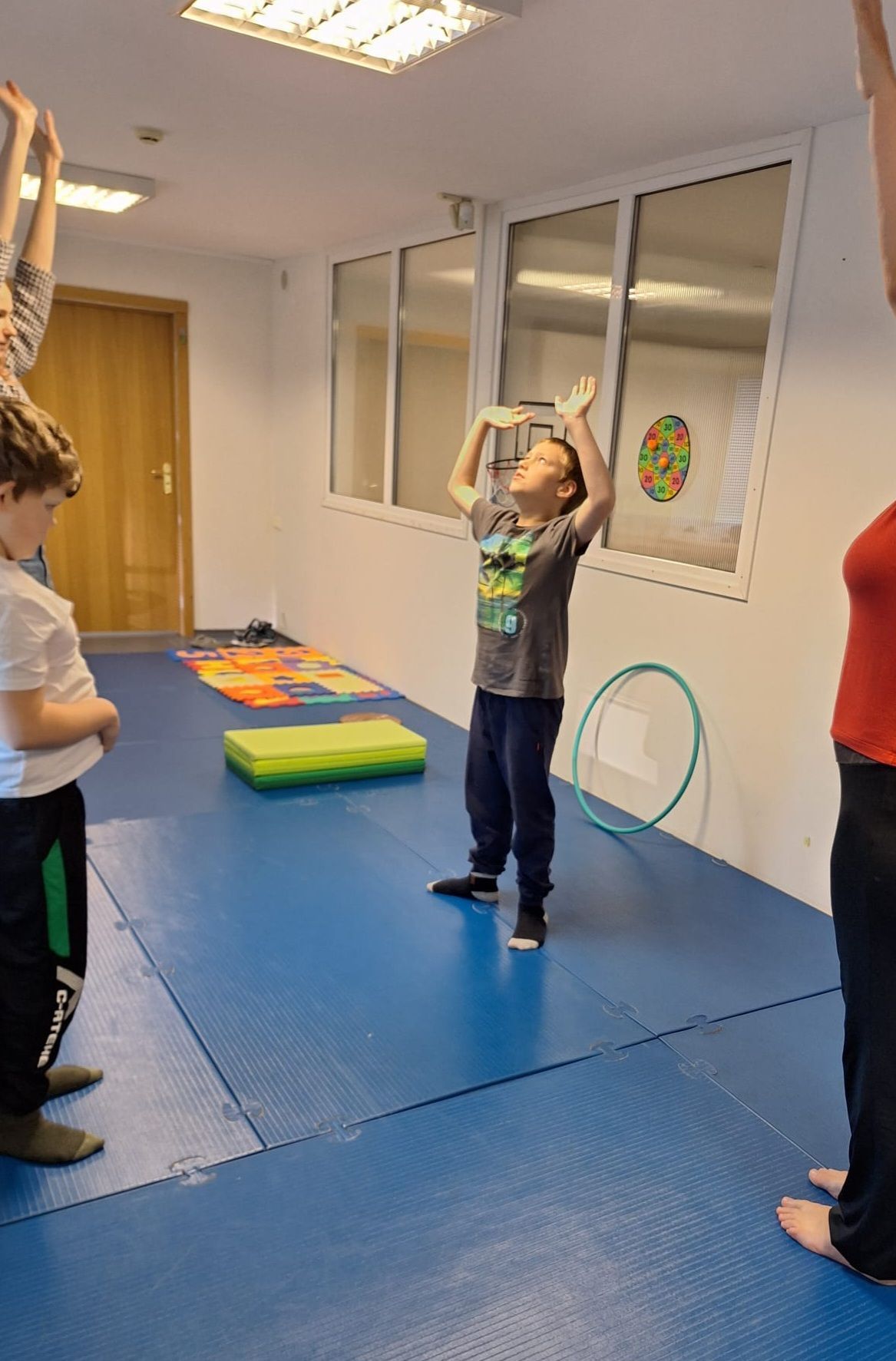 People exercise in a room with blue mats, a green foam pad, and a hula hoop. One child reaches their arms upward.