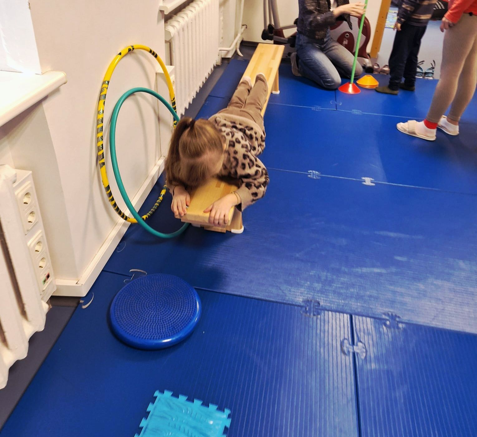 A child crawls on their belly across a low wooden bench in a gym, with hula hoops and sensory equipment nearby.