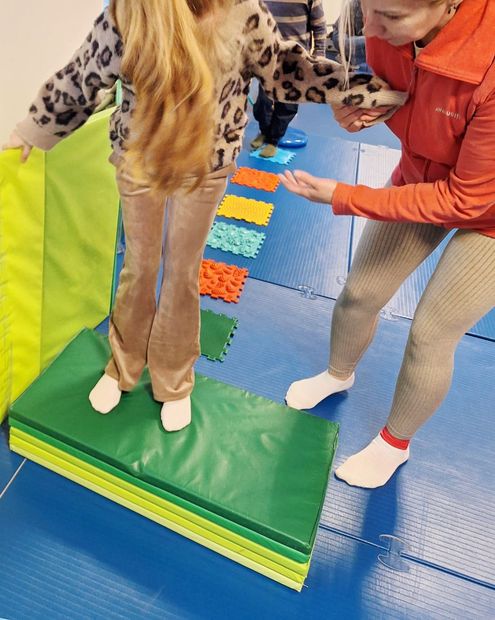 A child balancing on a raised green mat during a sensory activity with an adult guiding them.