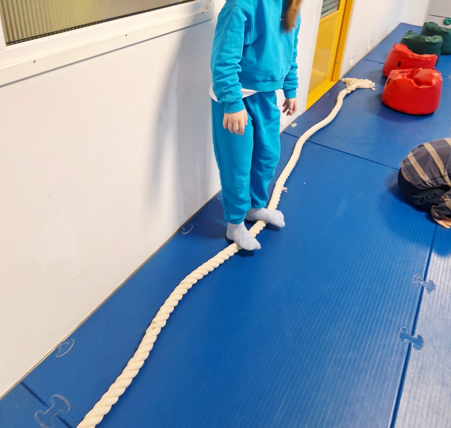 A person in a blue sweat suit balances on a rope laid across a blue gym mat, with colorful soft blocks nearby.