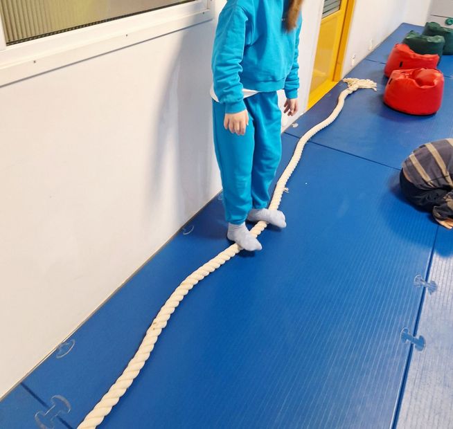 A person in a blue sweatsuit balances on a long white rope placed on a blue gym mat.