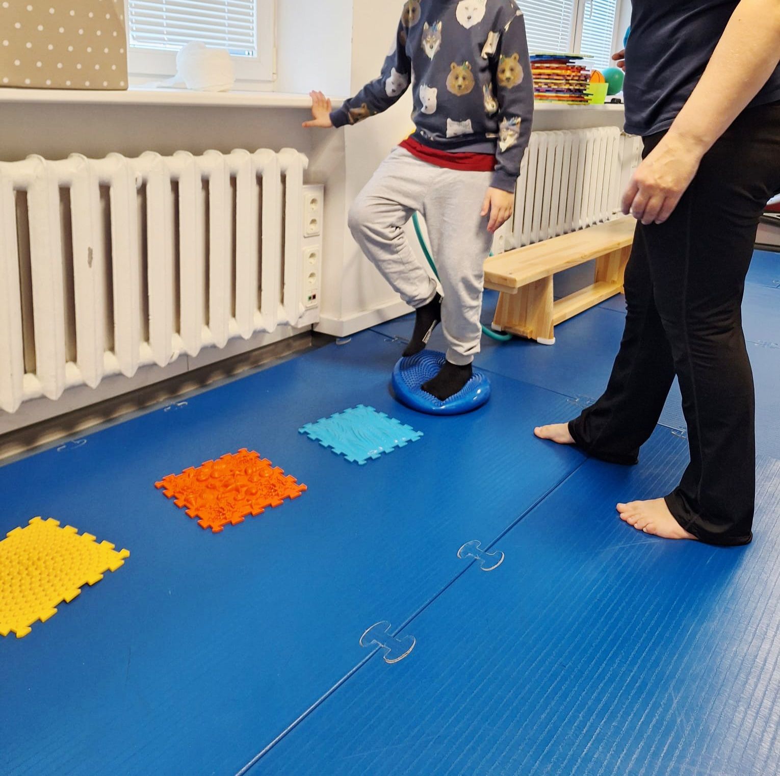 A person balancing on a blue vestibular disc in a therapy room, with colorful textured sensory mats on the blue floor.
