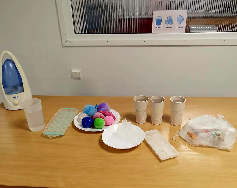 A table with a humidifier, cups, ice trays, colorful balls, and a bowl of ice for a sensory activity.