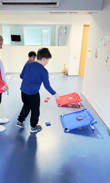 Children play a bean bag toss game with red and blue boards on a light-colored floor in a bright indoor room.