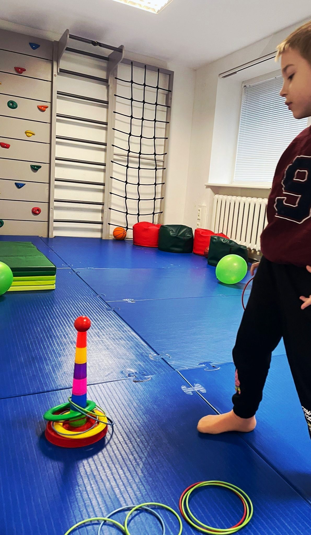 A person plays a ring toss game on a blue mat in a gym with a climbing wall and colorful beanbags in the background.