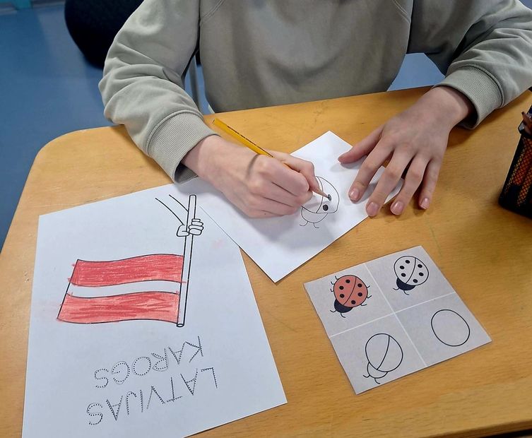 Hands hold a pencil, drawing on paper next to a picture of the Latvian flag and a sheet with ladybug illustrations.
