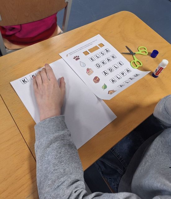 A student at a wooden desk works on a cut-and-paste literacy worksheet with scissors, glue, and letter tiles.