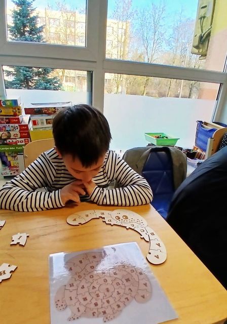 A person in a striped shirt sits at a wooden table assembling a wooden jigsaw puzzle shaped like a turtle.