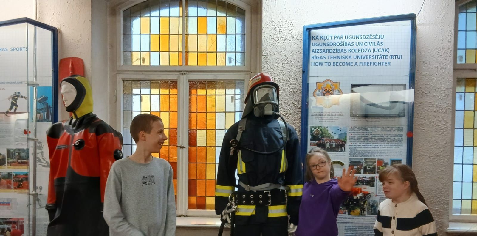 Three children stand near two firefighting gear mannequins in front of a stained-glass window in a museum exhibit.