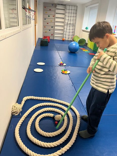 A child uses a green stick to guide a small ball through a rope spiral on a blue mat in a sensory gym.